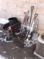 Photo showing the five shovels, red-handled gardening fork, black garbage can, and garden cart arranged outside against a brick wall with some snow on ground.