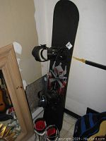 Full view of Dakine snowboard standing upright with LX black bindings attached, along with Ride snowboarding boots at base.