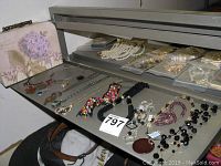 Photo showing all twelve costume jewelry necklaces spread out on a shelf, along with a small decorative jewelry box with floral design in the background.