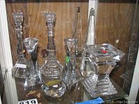 Front view of lot inside glass cabinet showing six Baccarat crystal items: two pairs of candlesticks, obelisk, and square candle holder.