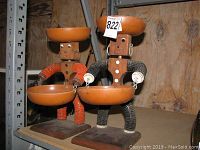Photo showing two Caribe folk art snack holder figurines, one with orange beaded limbs and one with black beaded limbs, both holding snack bowls and standing on wooden bases.