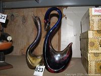 Two glass swan sculptures on a shelf showing both colors and forms from the front and side angles.