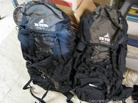 Two black Teton Sports backpacks side by side on floor, showing front with brand logos and external bungee cords.