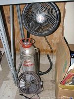 Two fans and fire extinguisher arranged on floor near shelving:Vornado mounted fan, Lasko fan in front, Pyrene extinguisher behind