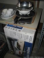 Photo of Waring Pro warming tray in original box alongside metal and plastic salad/pasta strainers and Farberware pancake pan with black handle on table.