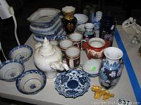 Photo of assorted bowls, vases, cups, teapot, dessert stand, gold dragon figurine, and pottery planters on a table.