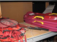 Photo shows badminton rackets and a red sled or toboggan stacked on a shelf. Image likely shows part of sporting goods items.