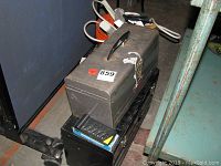 Closed metal toolbox on top of a black item, possibly another tool box or case.