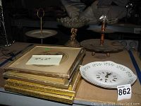 Lot arranged on a flat surface showing three vintage serving stands and stacks of decorative wall art frames with one ceramic wall clock plate included.