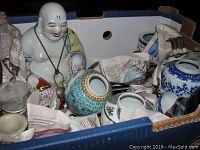 Box showing ceramic Buddha figurine, oriental bowls, and blue and white vases wrapped in newspaper.