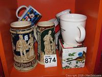 View of assorted steins and cups on orange shelf including two German steins, Starbucks cup, Mickey Mouse mug, box and additional white cups