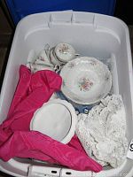 Wide shot of plastic bin containing several porcelain pieces: cups, plates, with floral pattern, some wrapped in bubble wrap, cloth liners present