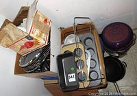 Photo showing assorted baking tins including muffin trays, loaf pan, round tins, various wire racks, cooking pans, and purple teapots stacked together in cardboard boxes.