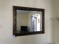 Front view of large square beveled mirror with bronze colored wooden frame hanging on wall, reflecting TV and doorway.