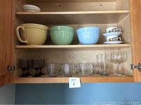 Open cabinet shelf showing large ceramic mixing bowls in beige, green, and blue, small floral teacups with saucers, various clear and brown stemmed glasses