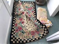 Large floral rug with black and beige checkered border displayed flat with two smaller folded rugs on a tiled floor.