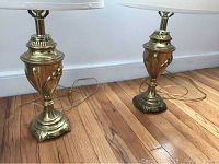 Pair of vintage brass urn-shaped lamps shown standing on wooden floor against a white wall. Each lamp includes a white drum lampshade and an electrical cord.