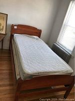 Full assembled twin bed frame made of solid maple wood with white mattress and box spring inside a room with wooden floor, plain white walls, and window with blinds.