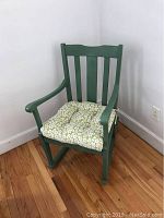 Front angled view of a small green wooden rocking chair with a white and green floral cushion on a wooden floor against a white wall.