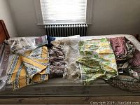 Full overview on bed showing multiple folded vintage linens and curtains including April Cornell and other French country style pieces.