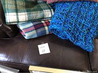 Photo showing three folded blankets on a brown leather surface: a green and cream plaid blanket, a red and blue plaid blanket, and a thick blue knit blanket.