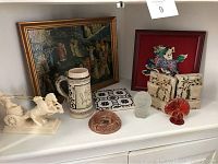 Shelf display showing the framed medieval style painting, carved horse chariot, stein, ashtray, frosted glass face sculpture, and carved stone fragments.
