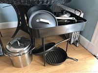 Wide view showing all pans and pots on a black cart and table surface, includes cast iron grill pan, Revere pot with lid, black non-stick pots and pans with white handles
