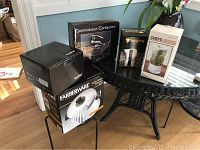 Five boxed kitchen items on a black glass table and metal chair. Boxes include Farberware pot, VonShef fondue set, corkscrew set, coffee maker and herb keeper.