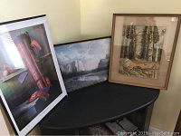 View showing three framed art prints leaning against the wall on a black table: a colorful Sid Willis golf print, a mountain landscape print, and a birch tree watercolor print.