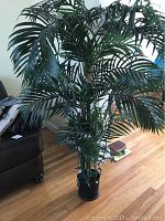 Front view of large faux bamboo tree showing green leaves, bamboo-style trunk, and black pot base on wood floor with nearby furniture.