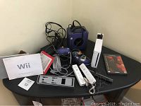 A side view of the Nintendo GameCube and Wii consoles with accessories arranged on a black table including remotes, cables, manuals, and a game case.