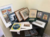 Overview of various framed and matted prints and artwork laid out on a piece of furniture. Includes city-themed prints with visible city names such as New York and visible frames of different styles.