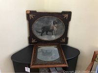 Carved wood framed dog art with image of a dog in an oval-shaped inset and carved corner decorations, placed on a table behind a carved wood framed rectangular mirror.