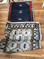 Two rugs displayed on wooden floor: one laid flat showing blue field with camel motifs, the other partially folded showing cream background with floral and heart designs.