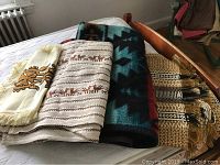 Four folded textiles displayed: a cream-colored runner with brown motifs and fringes, a white runner with brown horizontal patterns, a thick Southwest patterned blanket in turquoise and burgundy, and a woven striped textile with fringes.