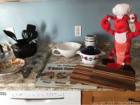 Full view of kitchen items on countertop showing cutting boards, bowls, utensil holder with utensils, lobster plush toy, decorative mat and stainless steel rack.