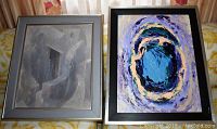 Two framed modern art paintings, one abstract with swirling blue/purple colors and the other with muted gray tones, both on fabric surface.
