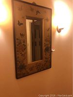 Full view of vertical rectangular wall mirror showing floral and butterfly designs on the wooden frame, mounted on a beige wall with a sconce lighting nearby.