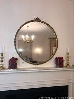 Full view of the round goldtone framed mirror hanging above a mantel with two candle holders and berry decorations nearby.