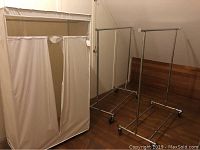 Photo showing zippered fabric wardrobe along with two metal rolling garment racks on casters.