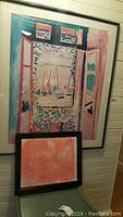 Large framed art print showing a colorful stylized window opening to sailboats on water, with smaller coral/orange mandala print leaning against the wall below.