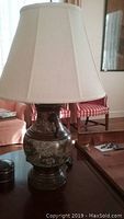 Metal lamp with beige fabric shade sitting on wooden container with brass hardware, alongside ceramic container and framed art piece.