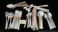 Full arrangement of most flatware pieces laid out on a dark surface, including teaspoons, soup spoons, knives, oyster forks, dinner forks, demitasse spoons, salad forks, and serving spoons.