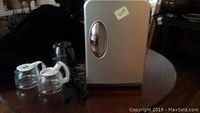 Closed silver mini desktop refrigerator with three glass coffee pot carafes and a few black coat hangers on a round wooden table.