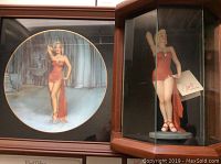 Photo of the Marilyn Monroe collectible plate displayed next to the glass and wooden display case containing the figurine.