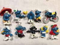Ten Smurfs figurines with various poses, colors, and accessories including one on a bicycle, arranged on a white fabric background.
