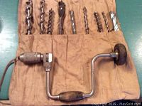 Photo showing manual brace hand drill and assortment of drill bits in cloth holder laid out on a green surface.