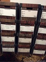 Front view of nine stacked Bankers Boxes showing labeled fronts with notes such as 'Picks - Xmas Short' and 'Pipe Cleaners'. Boxes have wood-like texture and black reinforced corners.