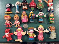 Top-down image showing a variety of approximately 17 small vintage plastic figurines including Miss Piggy, Mickey Mouse, clowns and others against a green surface with a US quarter for size reference