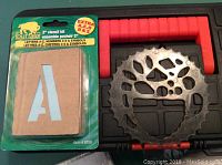 Photo of Buffalo brand 2-inch stencil kit containing letters A-Z, numbers 0-9, and symbols, still in original packaging, next to round black metal stencil.
